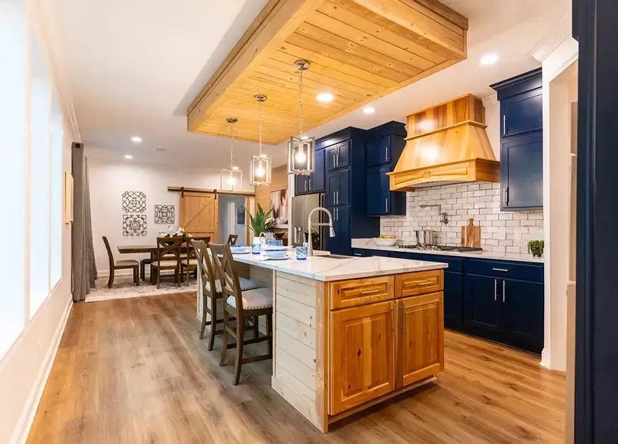 Modern kitchen with wooden island and ceiling details, blue cabinetry, stainless steel appliances, white brick backsplash, and pendant lighting, extending into a dining area with wooden furniture.