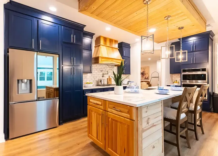 A mModern kitchen with blue cabinets, stainless steel appliances, and a wooden island. A wooden ceiling feature and pendant lights hang above the island, embodying the chic elegance you'd find in a Houston Shoreline Manufactured Home.dern kitchen with wooden cabinets, black appliances, and granite countertops. A vase with red tulips is on the counter. The living area has a sofa, round coffee table, and a rug.