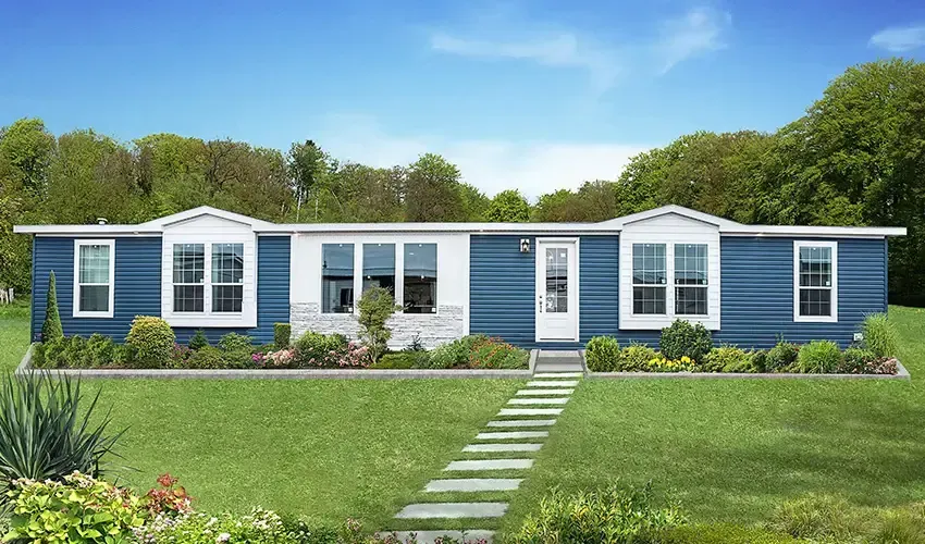 A single-story gray mobile home with white trim and a front porch, surrounded by a green lawn and plants, with a forested area in the background, offered by J Houston Properties Manufactured Homes.