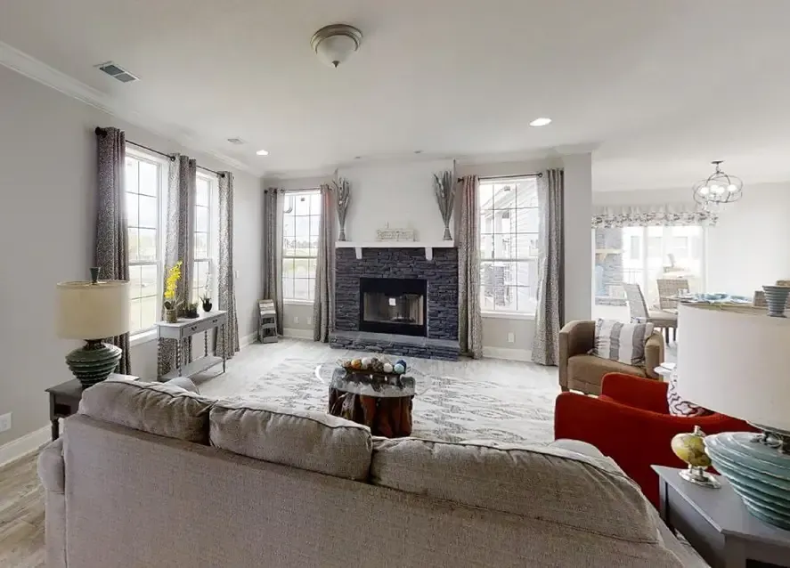 A modern, well-lit living room in a stylish Houston Nandia manufactured home, featuring a gray sofa, red chairs, and a stone fireplace. Large windows with drapes allow natural light to fill the room. The dining area is visible in the background.