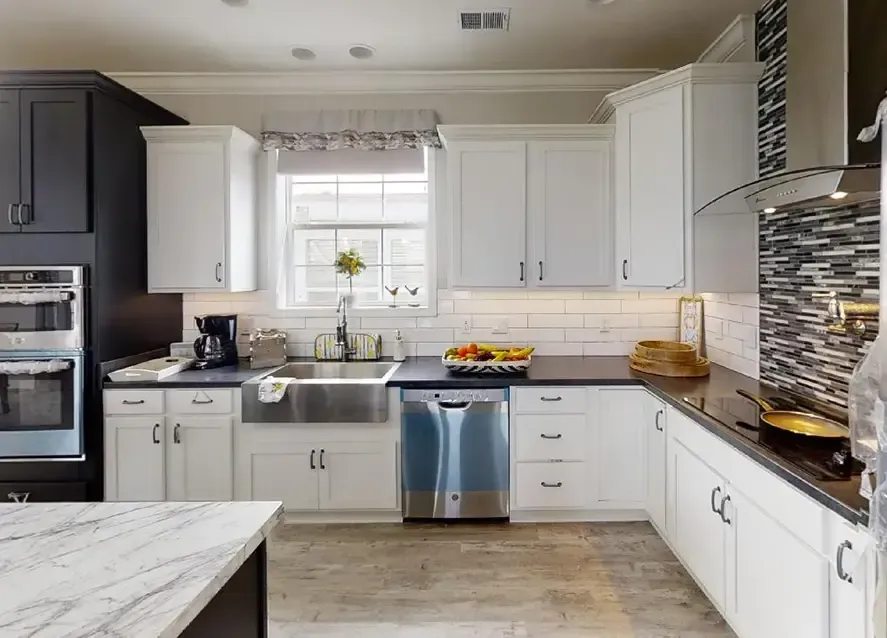 Modern kitchen in a Houston Nandia manufactured home featuring white cabinetry, stainless steel appliances, a farmhouse sink, and dark countertops. Tile backsplash behind the stove and items on counters including fruit and a coffee maker.