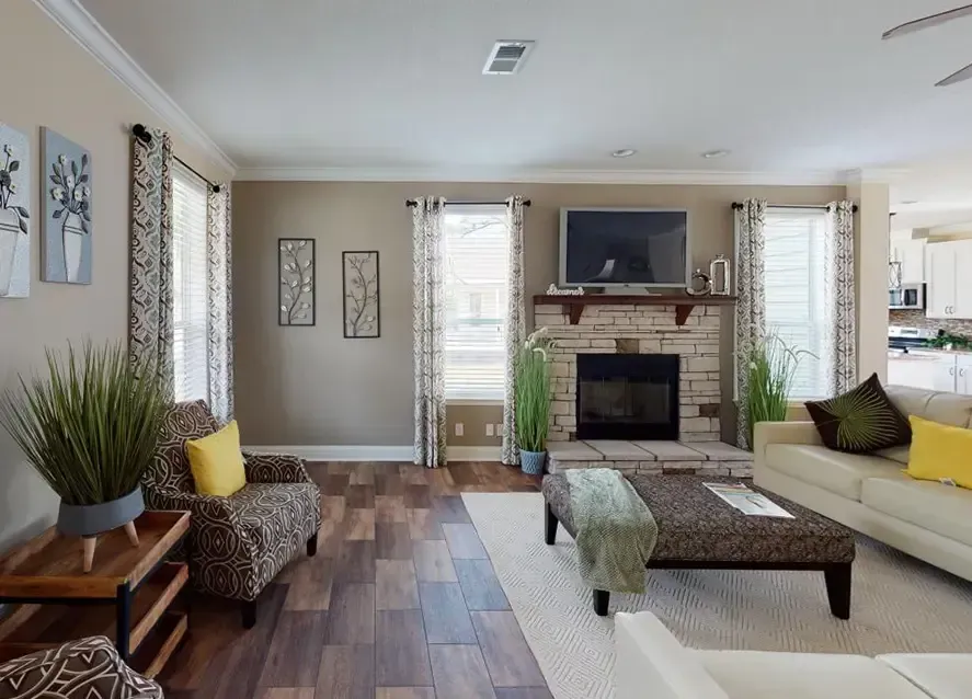 A modern living room in a Houston Lilly II manufactured home features a brick fireplace, flat-screen TV, and decor on the mantle. It boasts a beige sofa, patterned armchair, and coffee table on a hardwood floor, with large windows and curtains.