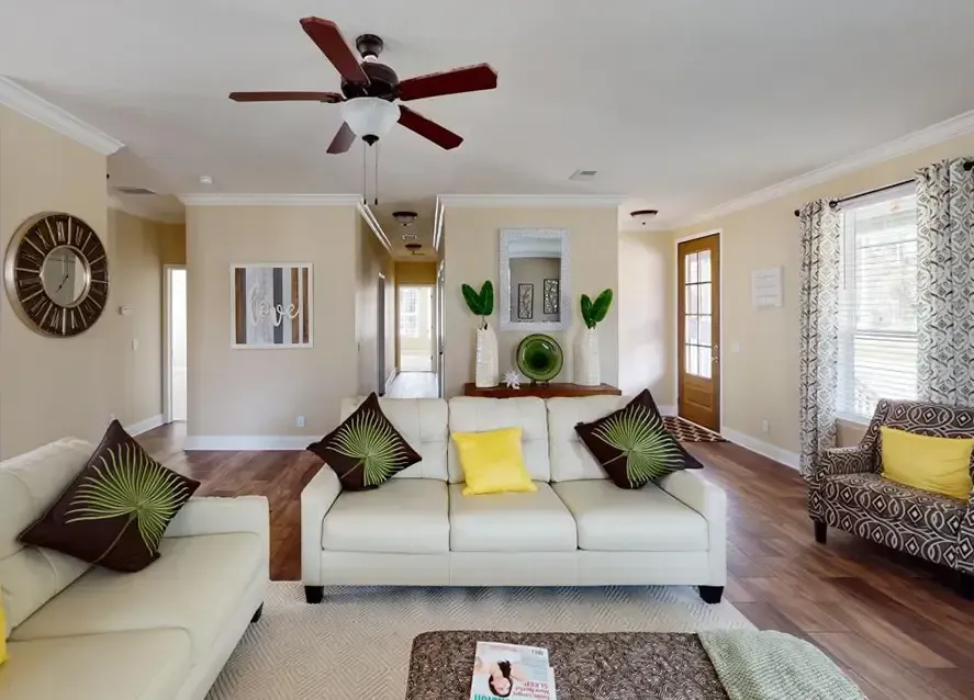 A tidy living room in a North Carolina manufactured home with beige walls, light-colored couches adorned with green and yellow pillows, a ceiling fan, and a patterned armchair. There's a magazine on the coffee table and art decor on the walls.