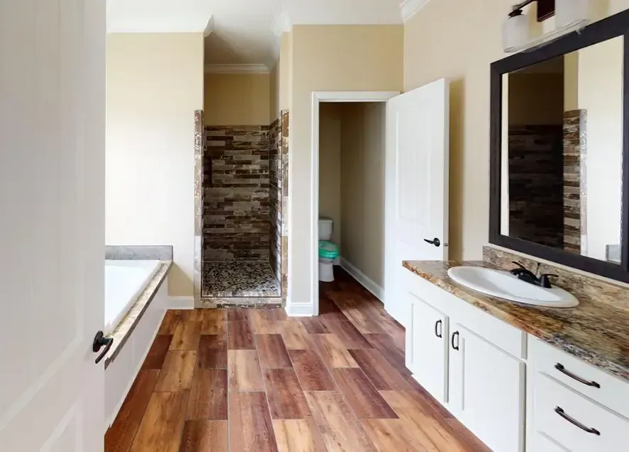 A bathroom in a Carolina Modular home features wooden floors, a large bathtub, a shower with stone-tiled walls, a toilet in a separate alcove, and a vanity with sink and mirror.