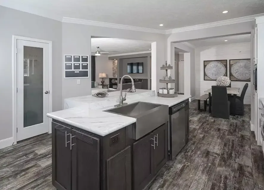 Modern kitchen with dark cabinets, marble countertops, a large stainless steel sink, and a built-in dishwasher. The kitchen opens to a dining area with dark furniture and monochrome wall art. This chic space is the perfect addition to any Houston Somerset modular home.