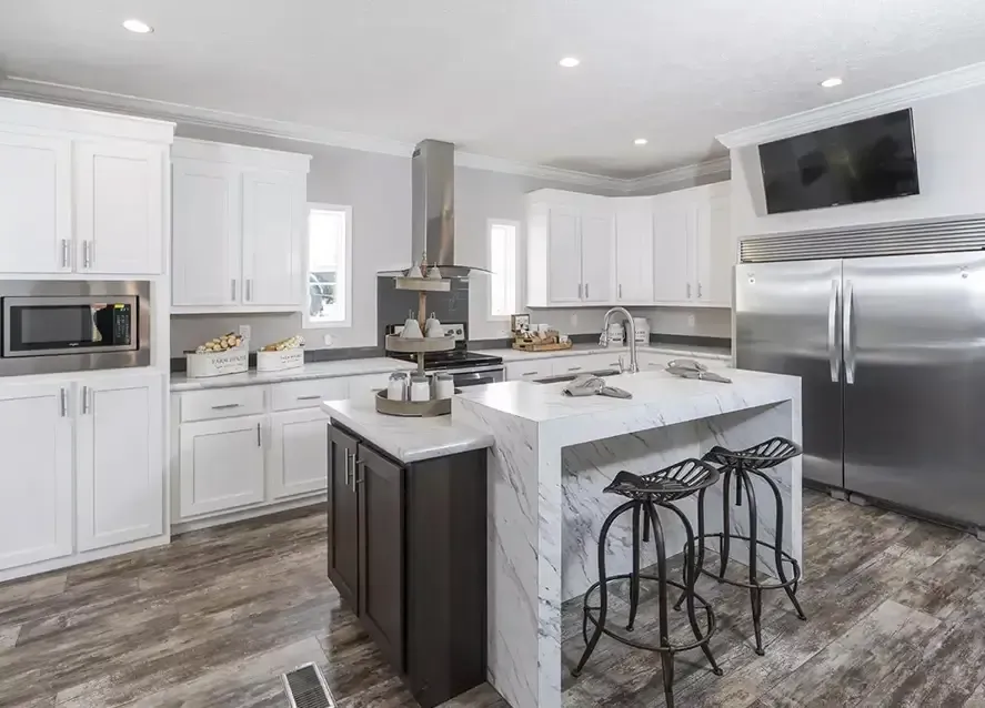 Modern kitchen in a Houston Somerset modular home features white cabinets, a marble island with two bar stools, stainless steel appliances including a large refrigerator, microwave, and wall-mounted TV.