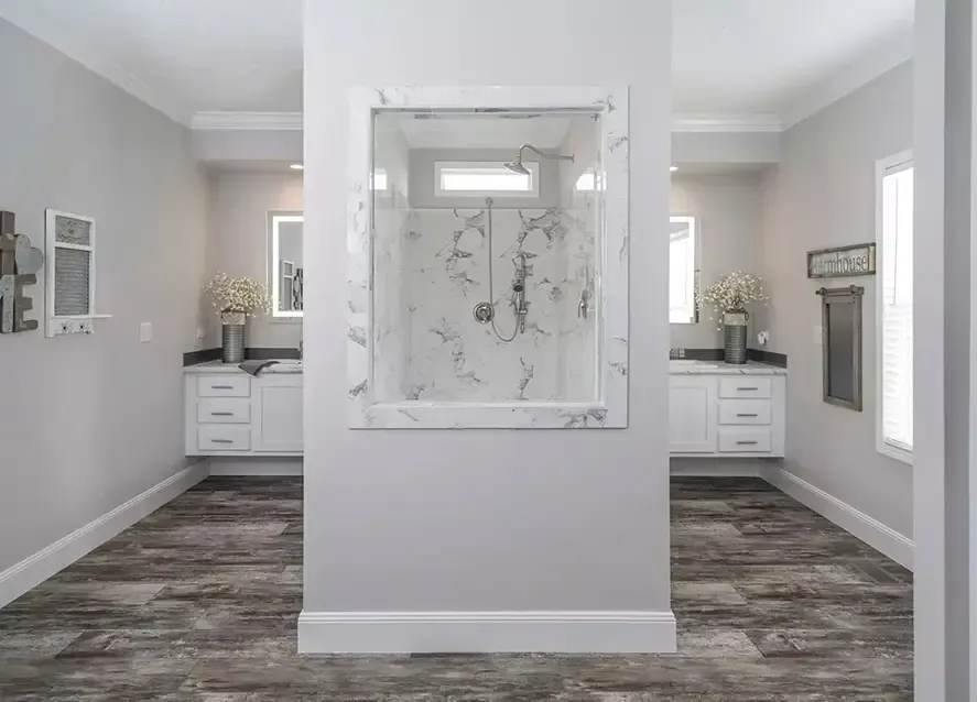 A modern bathroom in one of North Carolina's modular homes features dual vanities separated by a shower enclosure in the center. The floor boasts a wood-like texture, and the walls are painted light grey.