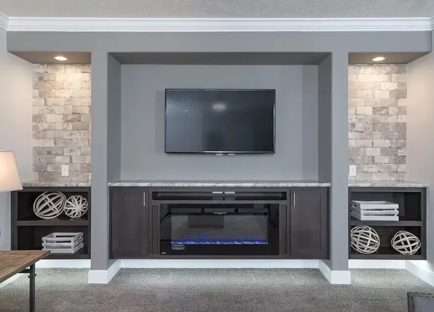 A modern living room setup in a Houston modular home features a wall-mounted TV above a fireplace. Shelves on either side contain decorative items and folded blankets, while the walls boast a stone-textured finish, epitomizing the charm you'd expect from a Somerset design.