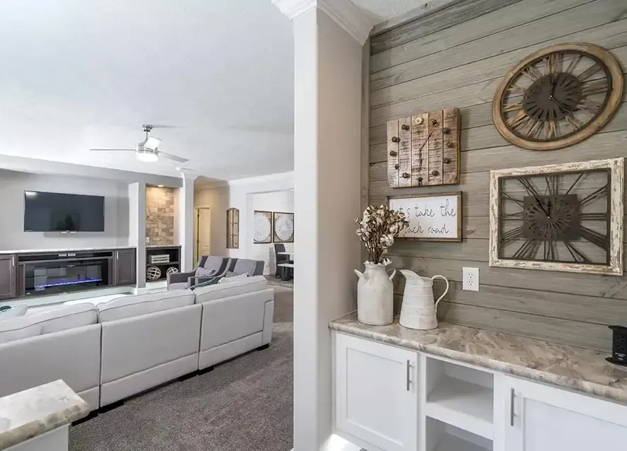 A modern living room in North Carolina modular homes features a white sectional sofa, wall-mounted TV, and fireplace. Timber accent wall with decorative clocks and wall art adds charm. White cabinets with marble countertops and decorative items complete the sophisticated look.