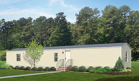 A beige mobile home from Houston Manufactured Homes, with a gray roof, front steps, and small bushes around its base. It is set on a grassy area in front of a dense forest.