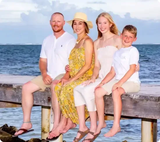 A family of four, including two adults and two children, seated on a wooden dock by the ocean. The adults and girl are dressed in light summer clothing, while the boy wears a white shirt and khaki shorts. Among them is the founder of JHouston Properties, enjoying a serene moment with loved ones.