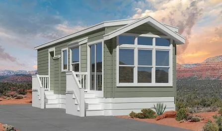 A small green and white manufactured home, from Houston Sedona Park Model Homes, with large front windows and steps leading to the front door, set against a desert landscape with rocky terrain and a cloudy sky.