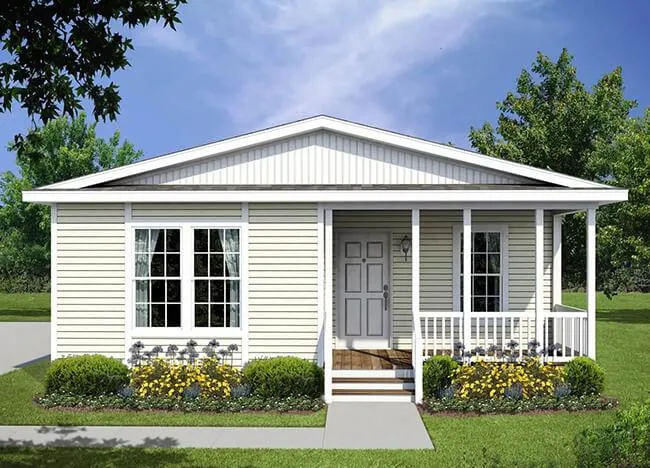 A small, single-story Houston modular home with light yellow siding, a front porch, and well-maintained shrubbery and flowers in the front yard, set against a backdrop of trees and a clear sky.