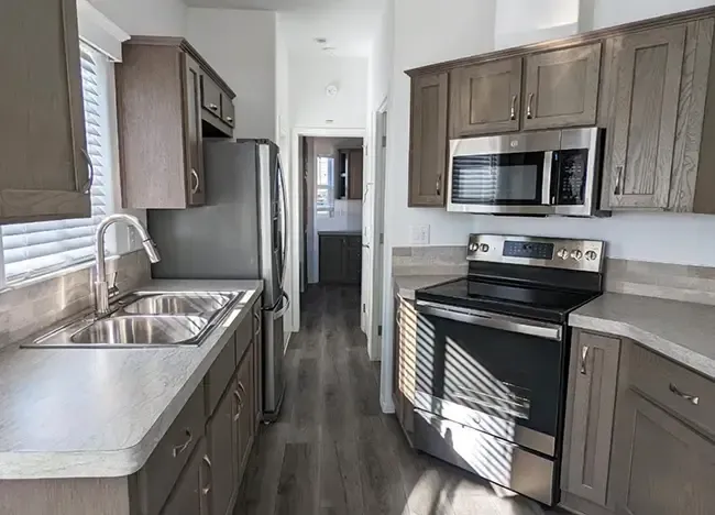 A modern kitchen with stainless steel appliances, including a refrigerator, stove, microwave, and double sink. Wood cabinets and light-colored countertops complement the room.