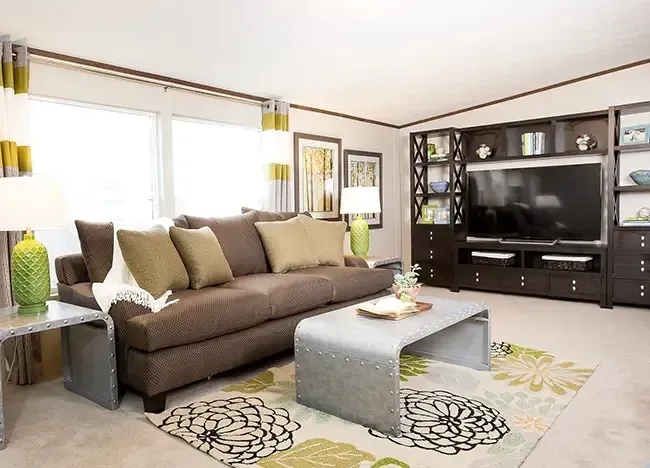 A living room with a brown sofa, gray metal table, large TV on a dark wood media unit, patterned rug, and two lamps on metal side tables. Decor includes green vases, framed artwork, and various trinkets.