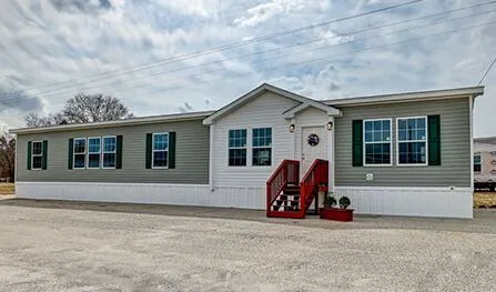 A single-story Houston ELK manufactured home with gray siding and white trim, featuring a red staircase leading to the entrance and a small wreath on the door. The surrounding area is a gravel driveway.