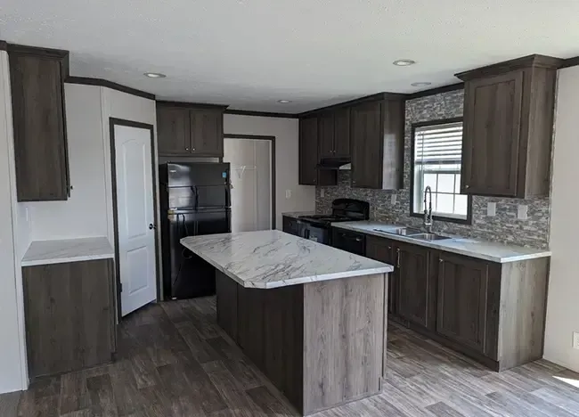 A modern kitchen in Houston Manufactured Homes featuring dark wood cabinets, marble countertops, a central island, stainless steel sink, black refrigerator, and stove. A door is visible on the left, and the floor has wood-like tiles.