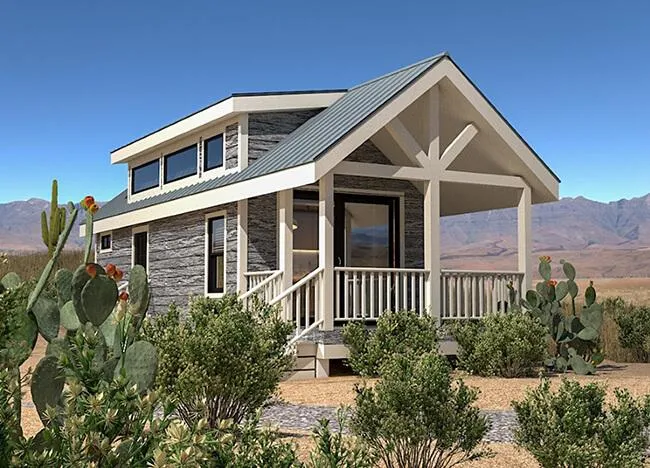 A small, modern cabin with a front porch sits in a desert landscape, surrounded by cacti and bushes, with mountains visible in the background.