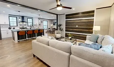 Open-concept living area with white sectional sofa, modern kitchen with island and bar stools, dining table, and wood accent wall. Ceiling fan is visible. Light wood flooring throughout.