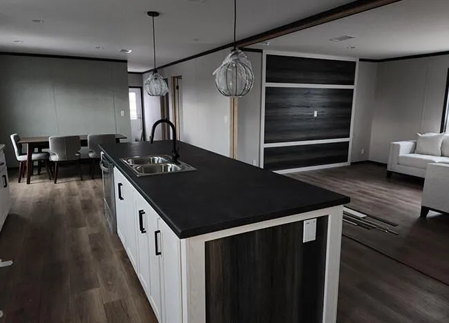 Modern kitchen and living area in a modular home, featuring a large island, black countertop, stainless steel sink, and pendant lights. Adjacent dining area with cushioned chairs, wood flooring, and minimalist decor.