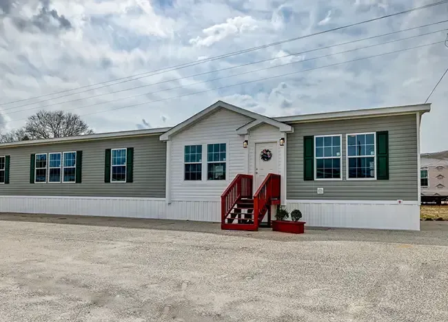 A single-story manufactured home with gray siding, green window shutters, and a red staircase leading to the entrance, set against a partly cloudy sky.