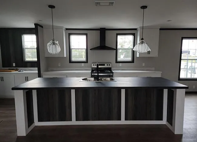 A modern kitchen features a central island with a matte black countertop, two pendant lights above, a stove with a range hood, and two windows on either side.