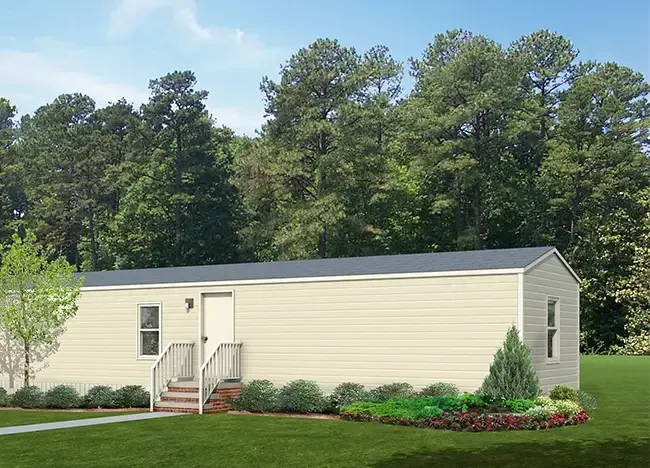 Single-story modular home with beige siding, a small porch with steps, and neatly landscaped surroundings. Tall trees stand behind the house.