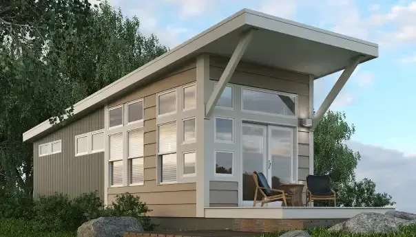 A small, modern Park Model RV home in North Carolina features a slanted roof, large windows, and a wooden deck with two chairs and a table, all surrounded by lush greenery.