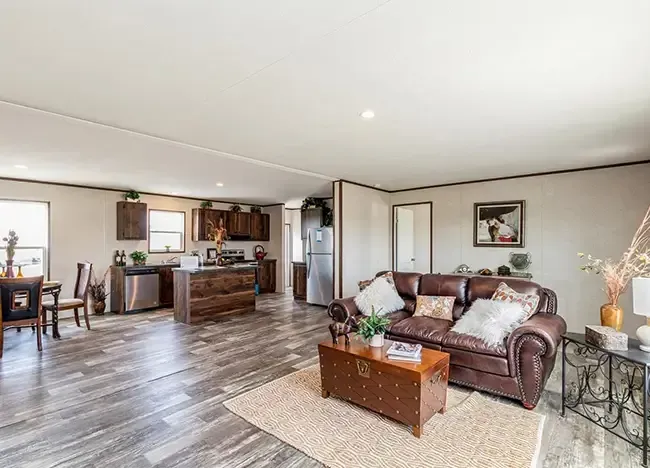 Open-concept living space with a brown leather sofa, a coffee table, a dining area, and a kitchen. The room has wooden flooring and simple decor with wall art and plants. Ideal for those looking at the charm of manufactured homes without compromising on style.