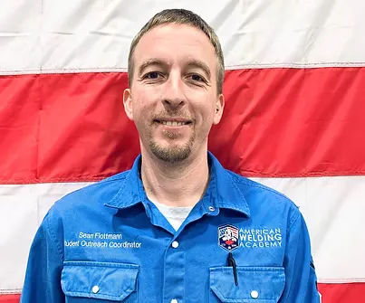 Man in blue uniform with “American Welding Academy” logo, standing in front of flag.