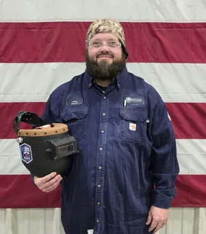 Man in dark uniform holding a decorated welding helmet, standing before a flag.