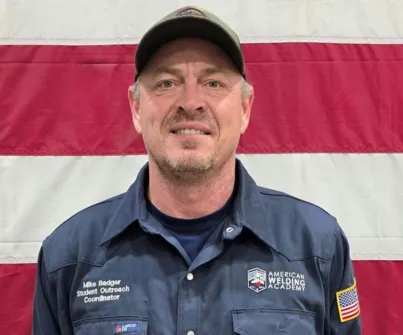 Man in blue uniform with “American Welding Academy” logo, standing in front of flag.