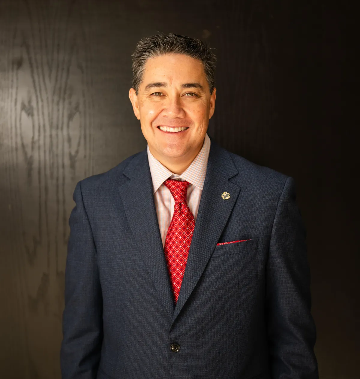 Professional headshot of smiling man in navy suit and red tie with dark background