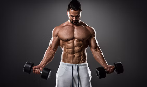 A muscular man in gray shorts holds a dumbbell in each hand, standing against a dark background and looking down at the weights.
