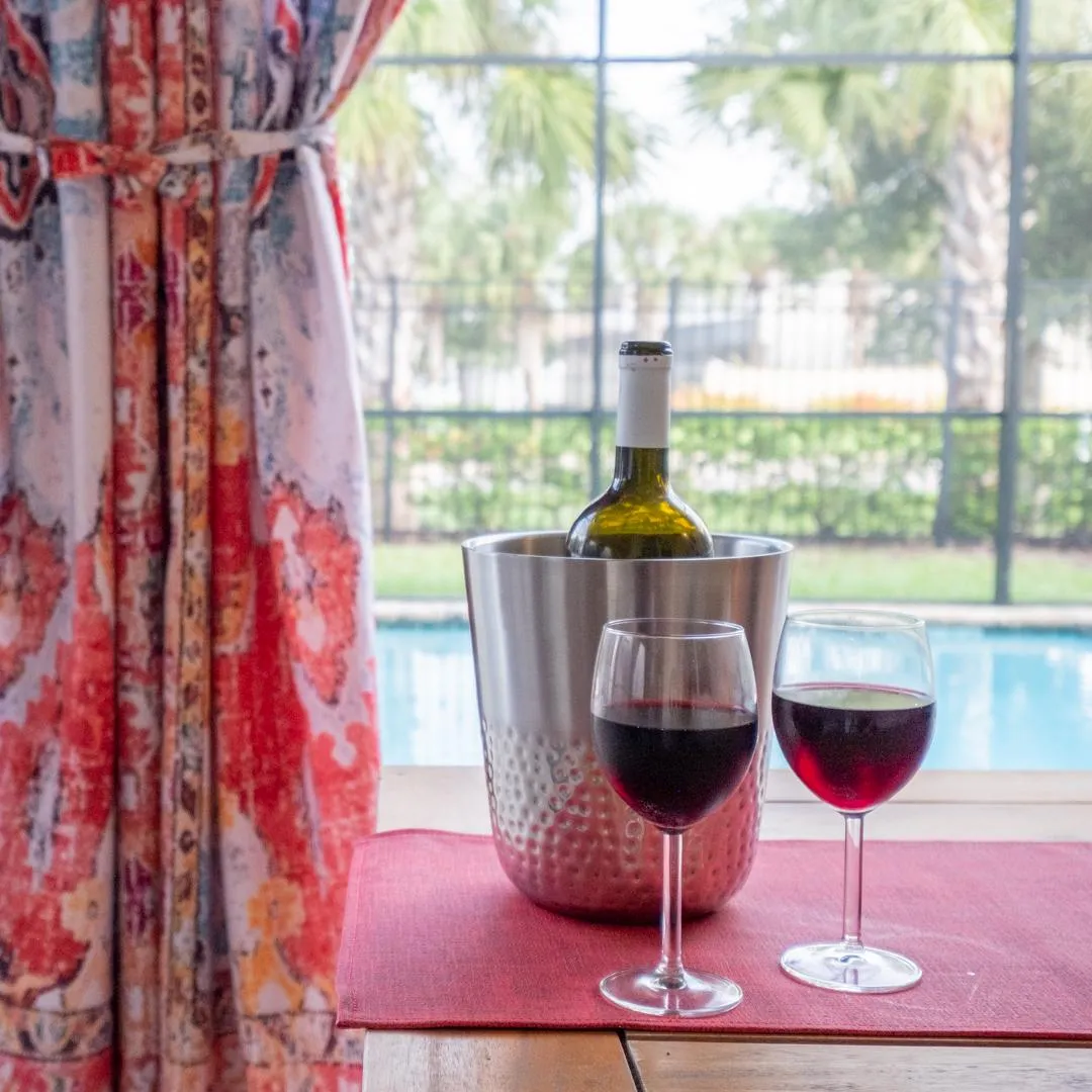 Bottle of red wine and two filled wine glasses in al fresco dining area overlooking pool and palm trees