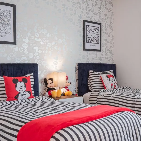 Mickey and Minnie Mouse themed bedroom with two twin beds, black and white striped comforters, red accent blanket, and mouse throw pillow. Mickey Mouse wallpaper and artwork hangs on the wall. Plush Mickey and Minnie dolls are seated on the nightstand.