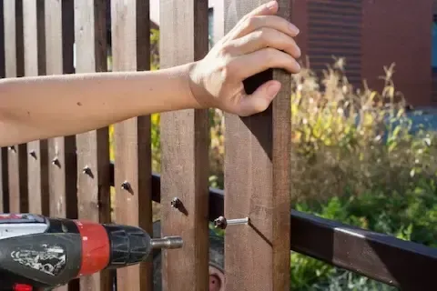 Person using a power drill to fasten a screw into a brown wooden fence post.