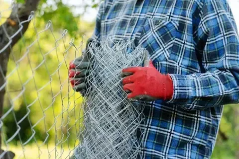 Person wearing plaid shirt and red gloves holding rolled chain-link fence outdoors.