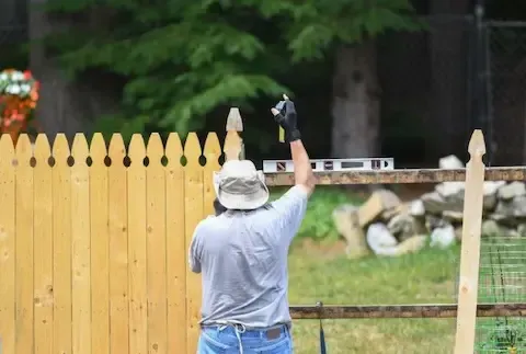 Man using level to build a wooden fence outdoors, holding a tool up, in front of greenery.