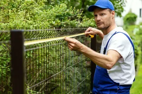 Professional fence contractor measuring metal garden fence with tape measure during installation in residential backyard.
