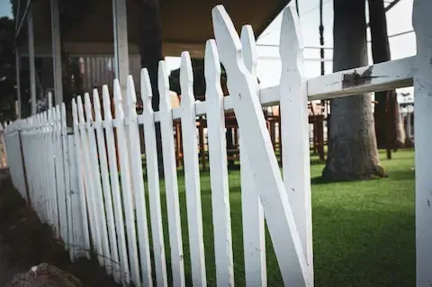 Damaged white wood picket fence leaning and broken in front yard.