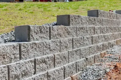 Concrete block retaining wall under construction with gravel base in a landscaped backyard.
