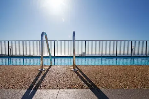 Black metal safety fence surrounding an outdoor swimming pool with clear blue water on a sunny day.