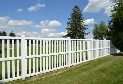 White vinyl privacy fence installed along a green residential lawn under a blue sky with scattered clouds.