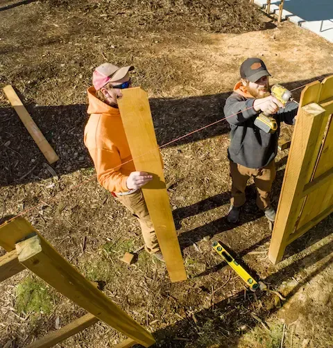 Two construction workers building wooden fence gate using drill and level on dirt site.