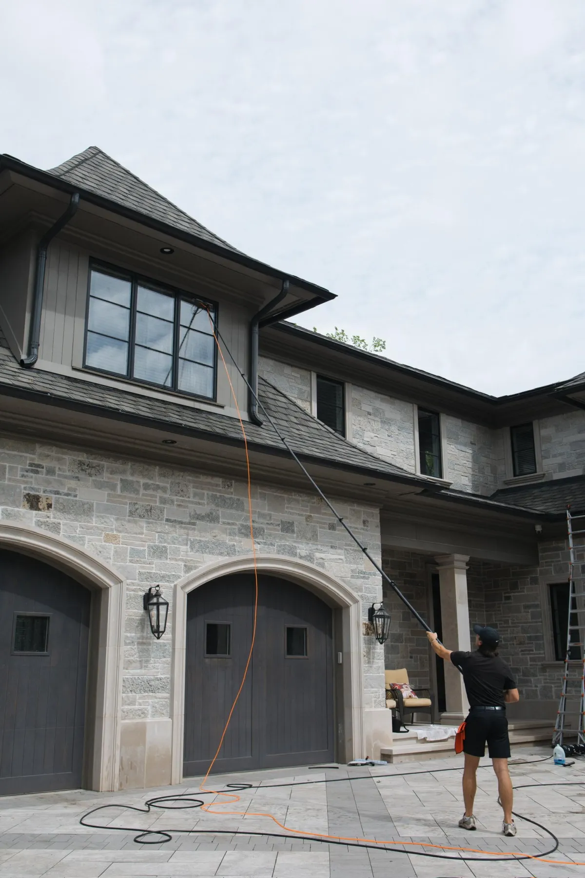 Professional window cleaner using water-fed pole system to clean second-story residential windows on luxury stone home