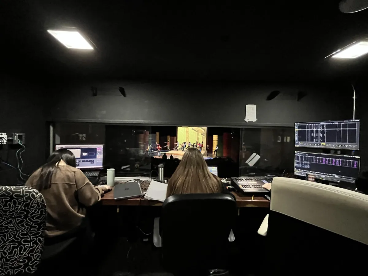 A view of a stage from the control booth, looking over the head of the stage manager. A rehearsal is happening on stage.