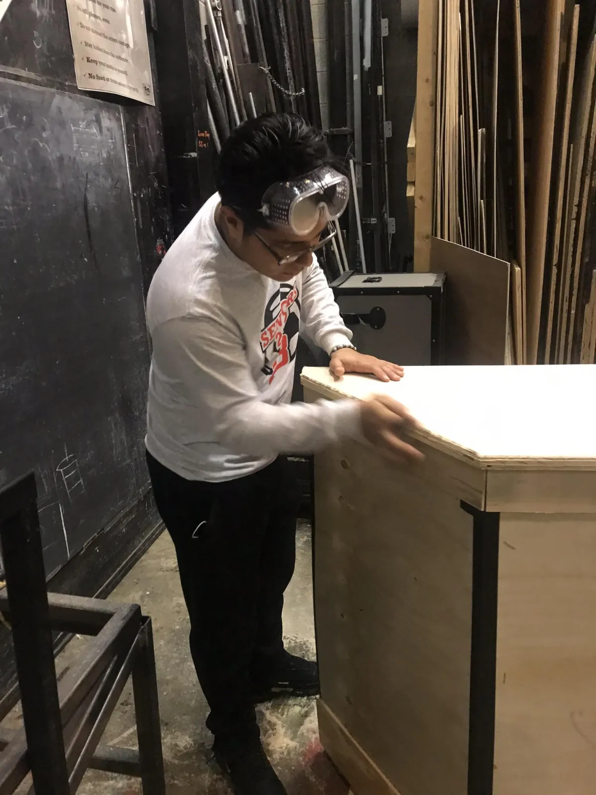 A student wearing safety goggles and over-glasses works carefully in a theater shop, sanding or assembling a wooden platform or set piece. The shop environment includes lumber, tools, and a dust collection system in the background, emphasizing a focus on safety and hands-on learning.