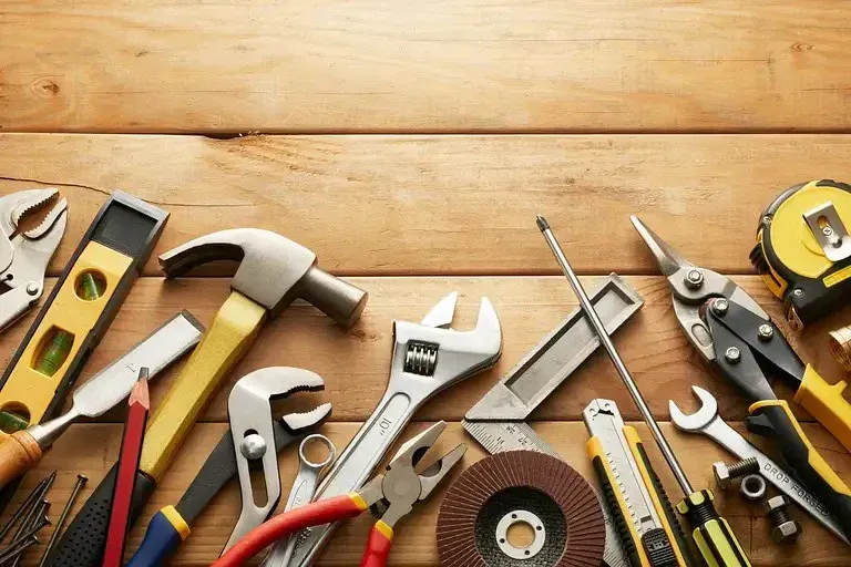 A variety of essential hand tools, including a hammer, adjustable wrench, pliers, screwdrivers, tape measure, utility knife, and sanding disc, on a wooden workbench.