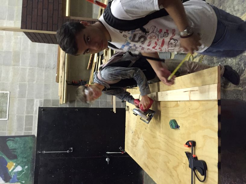 Two high school students working in a theater shop: one student is using a power drill to assemble wooden scenery on a workbench, while the other holds a tape measure. Both students are wearing safety goggles, and the workbench is equipped with clamps and woodworking tools. The background shows a well-organized shop space with lumber and storage shelves.